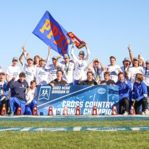 The Pomona-Pitzer men’s cross country team gather on the field in two rows as they raise hands in a number one gesture. One player at the center raises a large red trophy over his head and another player waves a blue flag with orange text that reads PPXC. Players in the front row hold up a blue sign with white text that reads 2023 NCAA Division III Cross Country National Champion.