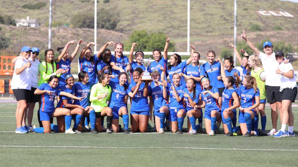 The Pomona-Pitzer women's soccer team gather with their coaches on the field in two rows while raising their hands triumphantly. A player at the center holds up a trophy. The team wears blue uniforms with white and orange accents.
