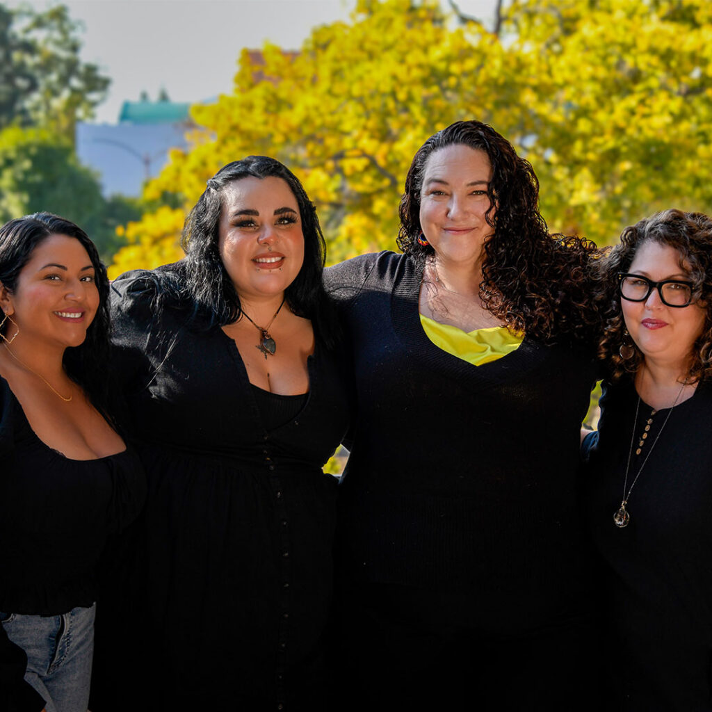 Jessica J. Chairez '13, Jack Contreras '22, Tricia Morgan '08, and Crystal Rodriguez '23 wear all black and stand side by side on a balcony overlooking Pitzer's campus.