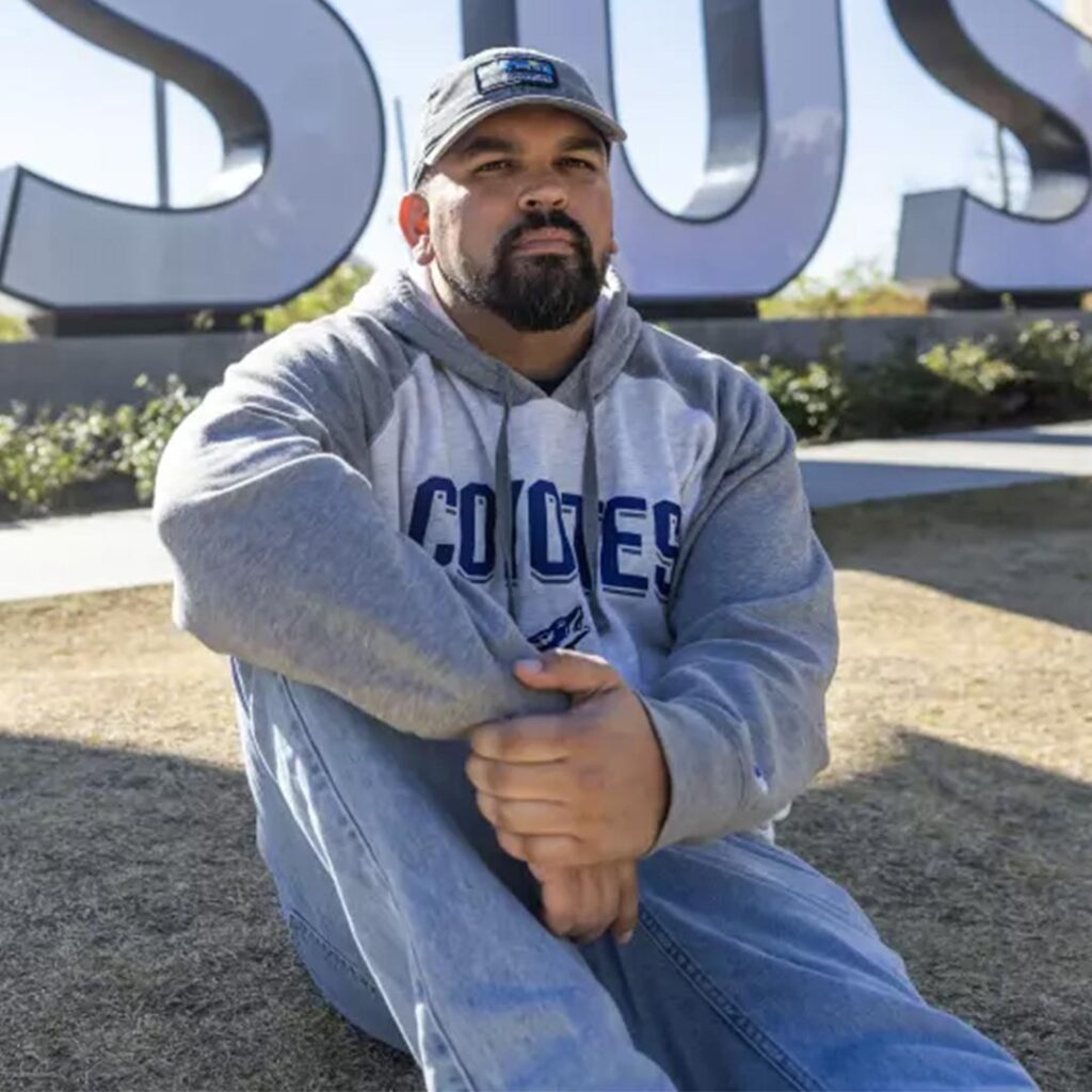 Michael Griggs sits on a grass hill with part of large CSUSB letter signs in the background. Griggs has a short black beard and wears a baseball cap, gray and white sweatshirt, and blue jeans.