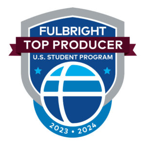 The Fulbright logo of white lines criss-crossing through a blue circle is overlayed on a blue badge with a gray outline and a dark burgundy ribbon cutting through the middle of the badge and above the circle. White text on the logo reads Fulbright Top Producer U.S. Student Program 2023 2024.