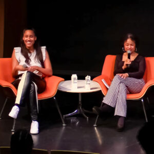 Anna Malaika Tubbs and Jan Barker Alexander sit side by side in orange chairs on a black stage. Barker Alexander holds up a mic while speaking to the audience.