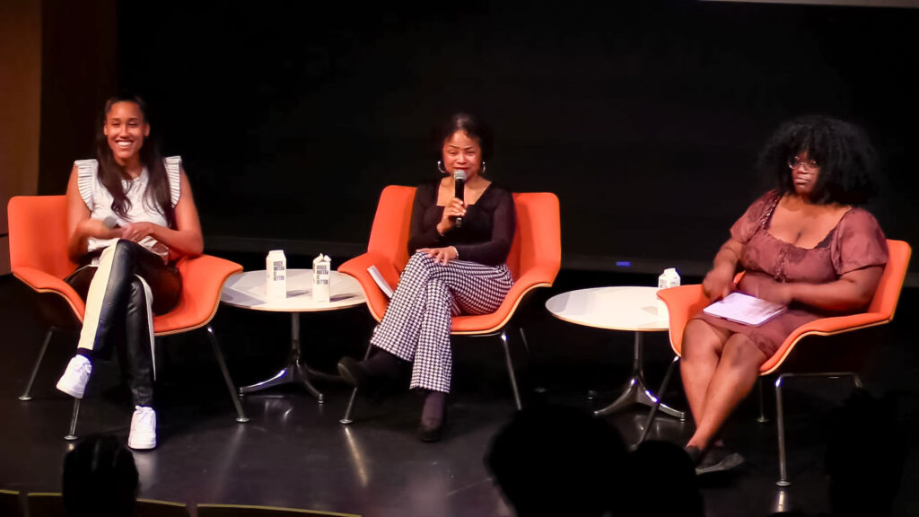 Anna Malaika Tubbs, Jan Barker Alexander, and Bee Joyner sit side by side in orange chairs on a black stage. Barker Alexander holds up a mic while speaking to the audience.