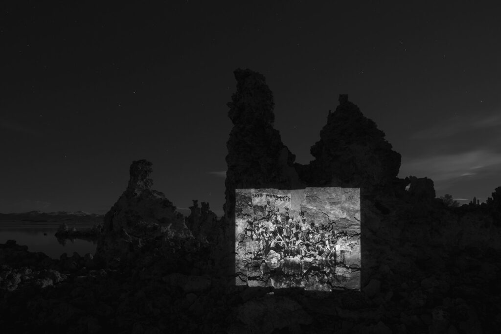 A black and white photo of a rock formation by Mono Lake at night. A photo of people gathered for a rehydration ceremony is projected on the rock formation.