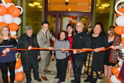 Interim President Tom Poon, Mayor of Ontario Paul Leon, and Pitzer and Ontario community members stand in front of the CASA Ontario building while cutting an orange ribbon.