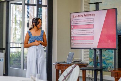 Pratya Poosala discusses her research while standing in front of a large screen. The screen shows a slide of her research with the title Brain-Mind Reflections at the top. Poosala has dark wavy hair pulled back and wears a blue top and white pants.