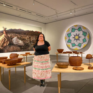Meranda Roberts stands in the middle of a ring of Cahuilla baskets sitting on curved tables. In the background are installations of a basket made of cans and a scenic photograph of a desertscape.