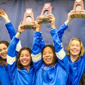 The Pomona-Pitzer women’s swim and dive team smile and hold up the NCAA trophies above their heads.