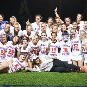 The Pomona-Pitzer women’s lacrosse team gathers for a group photo on the field and raises their fingers in a number one gesture. The team wears white uniforms with Pomona Pitzer and their individual numbers on the front in blue and orange font.