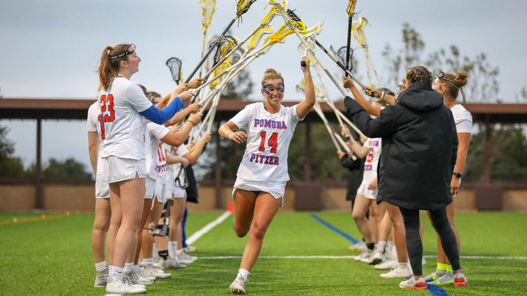 Carly Sullivan runs under a tunnel of lacrosse sticks held aloft by her teammates on her left and right. Sullivan has blond hair pulled back and wears the white lacrosse uniform with Pomona Pitzer 14 across the top.