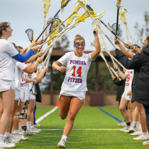 Carly Sullivan runs under a tunnel of lacrosse sticks held aloft by her teammates on her left and right. Sullivan has blond hair pulled back and wears the white lacrosse uniform with Pomona Pitzer 14 across the top.