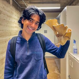 Tommy Shenoi holds up a large native oyster mushroom while standing in a lab. Shenoi has medium-length black hair and wears a backpack and a blue jacket.