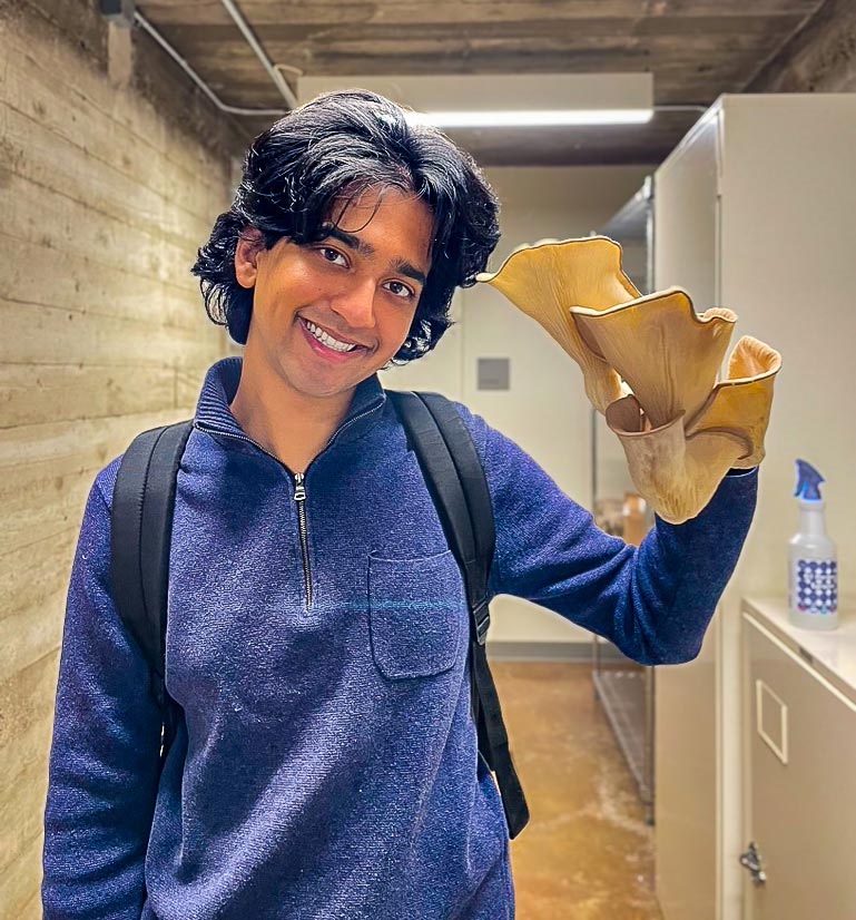 Tommy Shenoi holds up a large native oyster mushroom while standing in a lab. Shenoi has medium-length black hair and wears a backpack and a blue jacket.