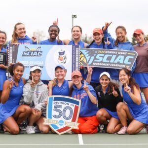 The Pomona-Pitzer women’s tennis team gathers on the court for a group photo. The team raises their fingers in a number one gesture and hold up signs for the SCIAC tournament championship.