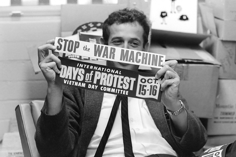 Edward Sampson, Associate Professor of Social Psychology, holds two bumper stickers: “International Days of Protest Vietnam Day Committee, Oct. 15–16” and “Stop the War Machine.” Taken September 16, 1965 in his office.