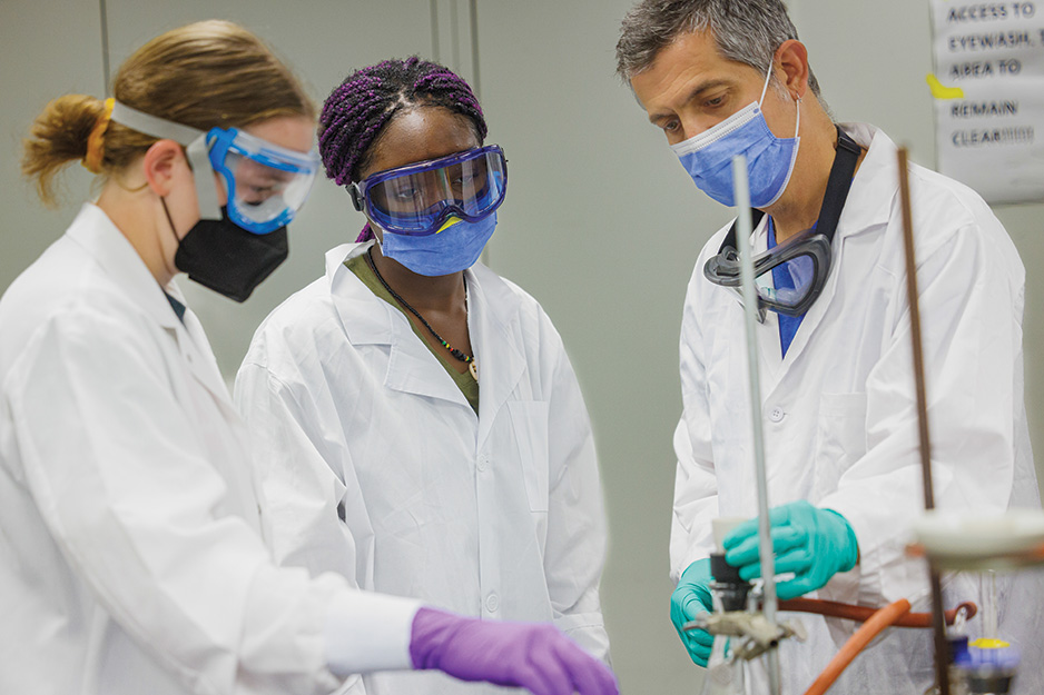 two students and a professor in white lab jackets and safety eyewear
