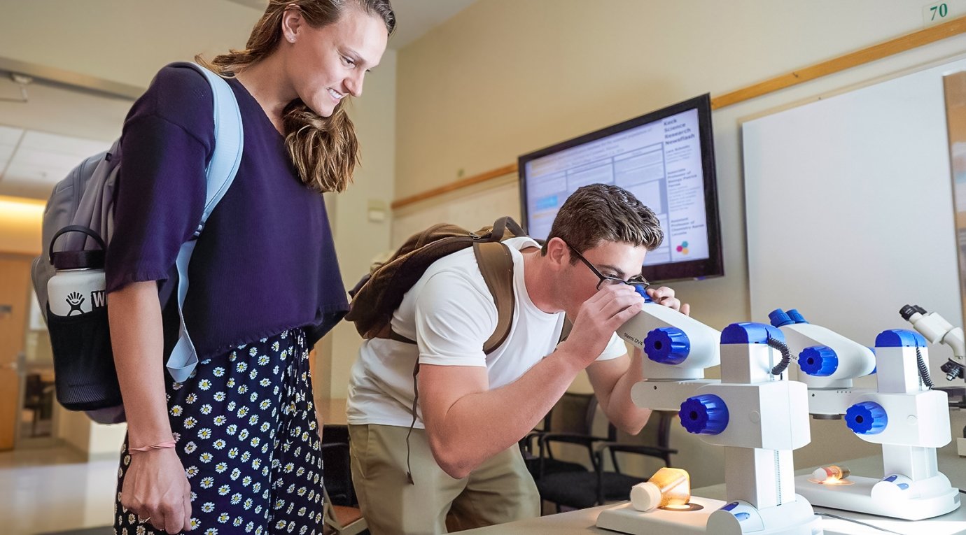 a student peers into a microscope while his classmate looks on