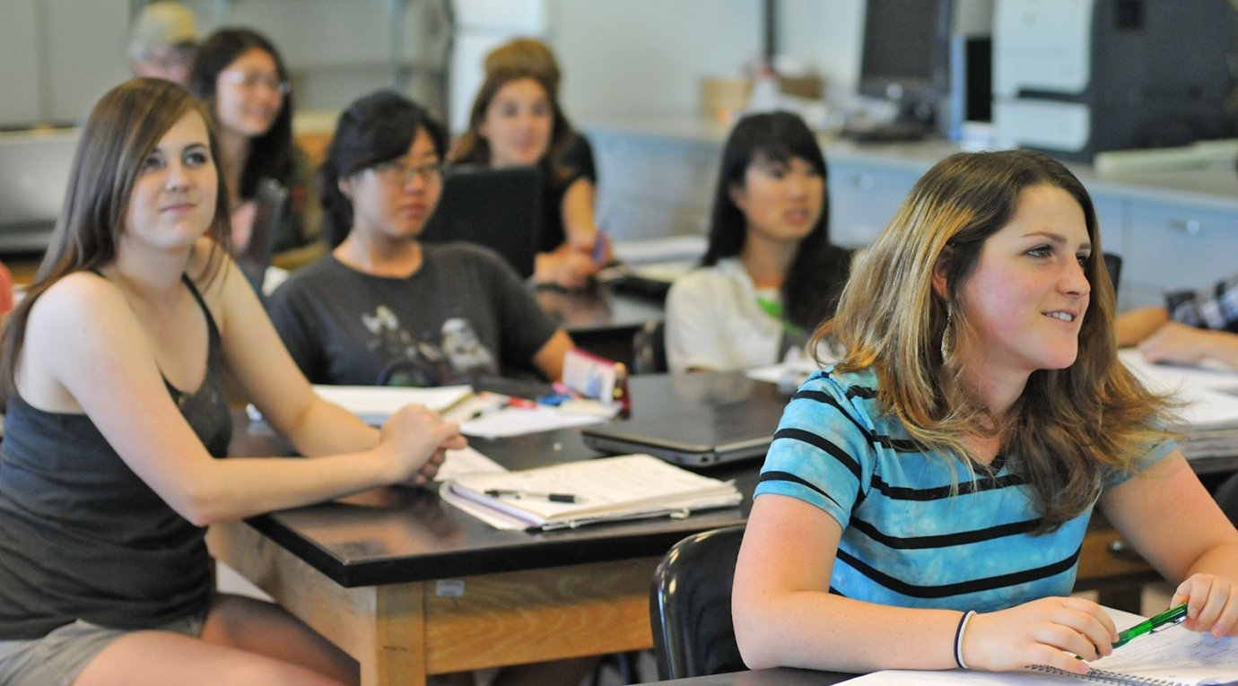 students sit at tables in a science classroom and watch the teacher lecture