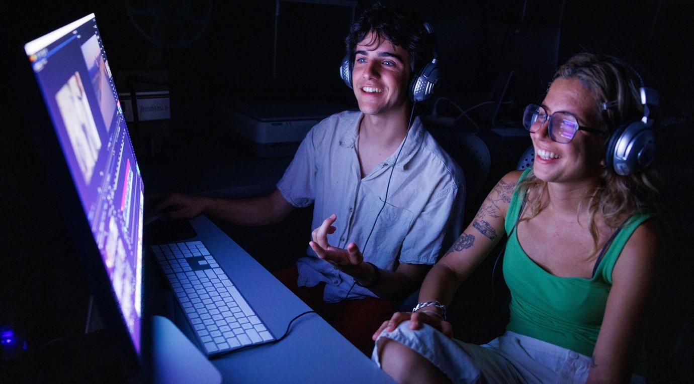 two students sit in a dark room in front of an computer screen in an editing bay