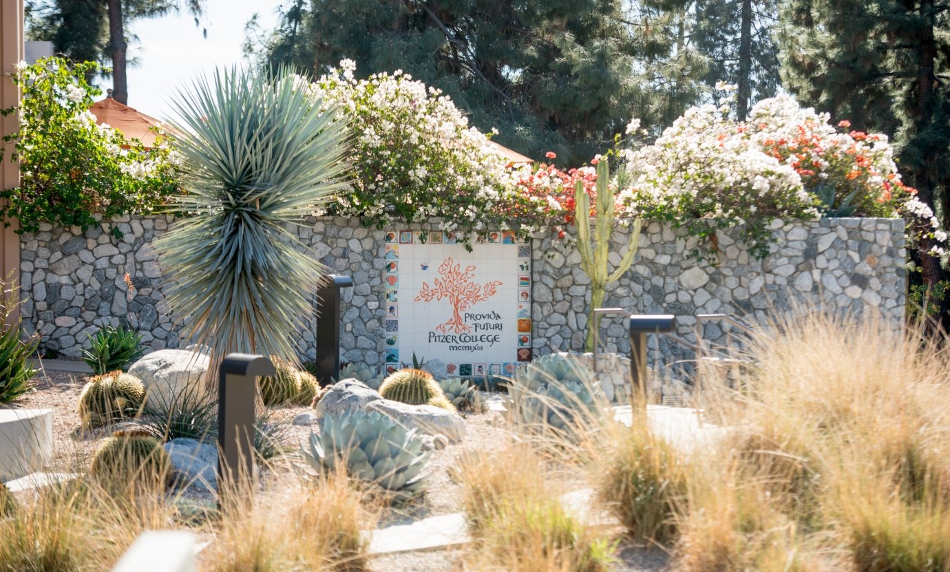 a desert landscape with a rock wall featuring sign with the words "Provida Future Pitzer College MCMIXIII"