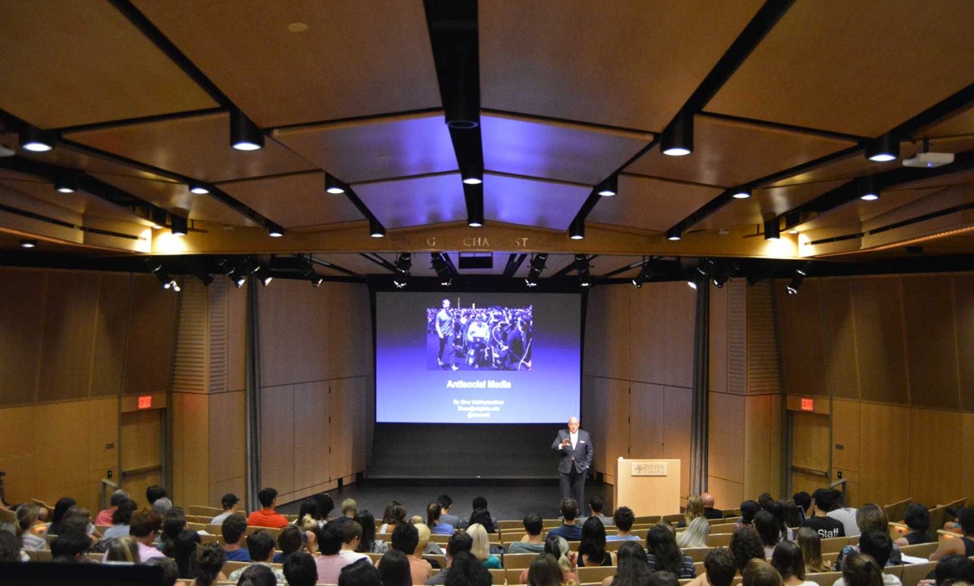 a presenter stands on stage in Benson Auditorium with a presentation on the screen behind him