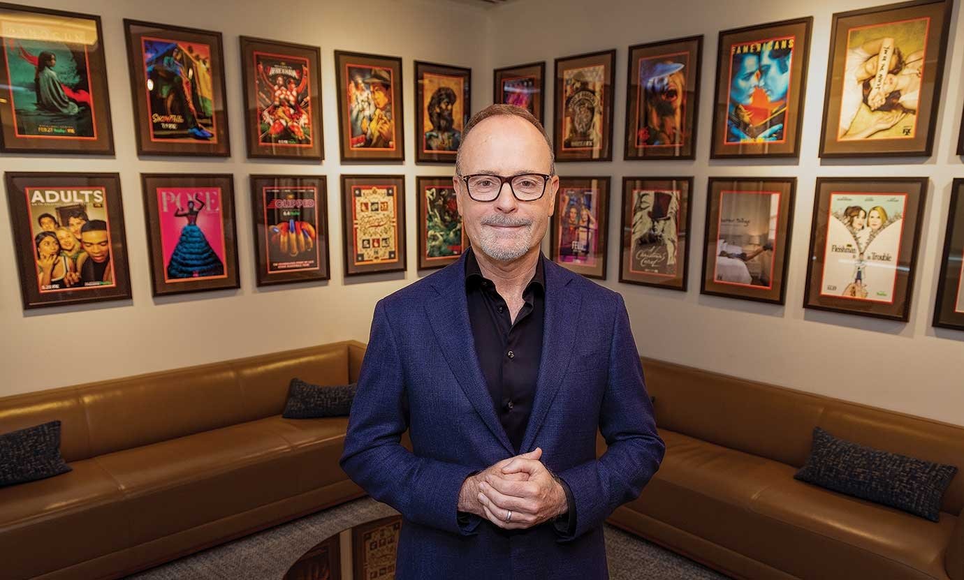 A man wearing a blue blazer poses in a room with several television show posters on the wall