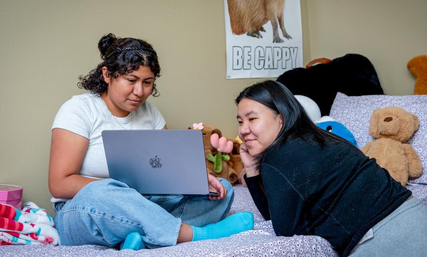 two students hang out on a bed in a dorm while looking at a computer