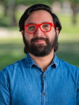 Man with dark hair and facial hair, wearing red glasses, and wearing blue-collared shirt, standing in front of greenery.