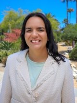 Mara Barbosa has long wavy black hair and wears a white blazer over a pale blue top while standing outdoors.
