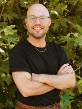 Man wearing glasses, a black shirt, and brown jeans crossing arms.