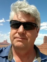 Man in blue shirt wearing black glasses with white short hair.