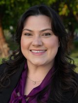 Woman in purple blouse wearing black jacket with curly black hair.