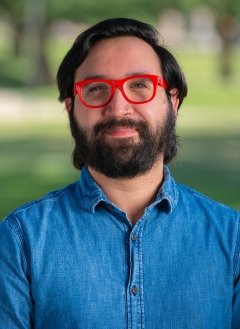 Man with dark hair and facial hair, wearing red glasses, and wearing blue-collared shirt, standing in front of greenery.