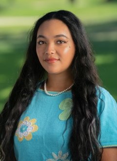 Woman with long hair, wearing a blue shirt, standing in front of greenery.