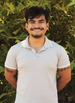 Man with white shirt and short black hair standing in front of bushes.