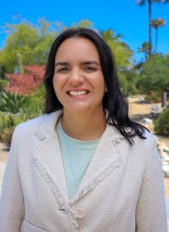 Mara Barbosa has long wavy black hair and wears a white blazer over a pale blue top while standing outdoors.