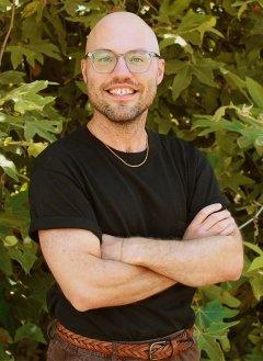 Man wearing glasses, a black shirt, and brown jeans crossing arms.