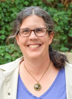 Woman in khaki coat and purple shirt wearing glasses with short brown hair.