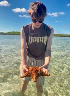 Man with brown short hair and brown tank top wearing glasses and holding a starfish in the ocean.
