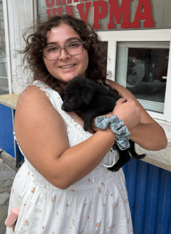 Woman in white dress wearing glasses with curling brown hair, holding a puppy in arms.