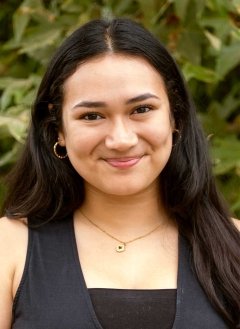 a student with long brown hair wears a black vest while standing in front of a plant on campus