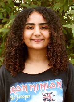 a student with curly long brown hair wears a iron minion tshirt while posing in front of a bush
