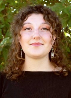 a student with long curly brown hair wears a black shirt while standing in front of a large bush