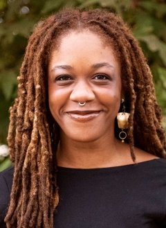 portrait of Chi Adi wearing a black top and long earrings, standing in front of an outdoor plant