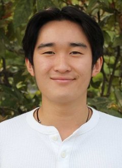 a student with short black hair wears a white henley shirt while standing in front of outdoor bushes