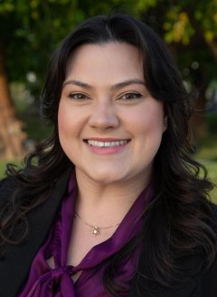 Woman in purple blouse wearing black jacket with curly black hair.