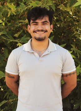 Man with white shirt and short black hair standing in front of bushes.
