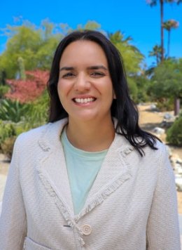 Mara Barbosa has long wavy black hair and wears a white blazer over a pale blue top while standing outdoors.