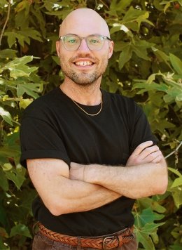 Man wearing glasses, a black shirt, and brown jeans crossing arms.