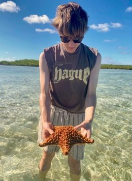 Man with brown short hair and brown tank top wearing glasses and holding a starfish in the ocean.